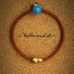 May include: A handcrafted bracelet featuring a red cord, a turquoise bead, and a gold-coloured bead. The text "-YoBracelet-" is written in black cursive. The bracelet is displayed on a textured, brown surface.