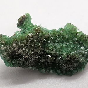 May include: A cluster of green fluorite crystals with dark grey and white inclusions. The crystals have a geometric shape and are clustered together, creating a textured appearance. The mineral specimen is set against a white background.