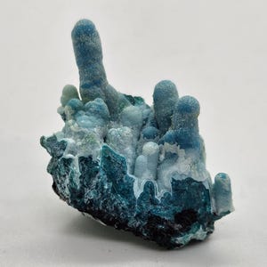 Könnte beinhalten: Nahaufnahme einer Mineralprobe mit einer Ansammlung von blauen, türkisfarbenen und weißen Formationen. Die strukturierte Oberfläche weist abgerundete und längliche Formen auf. Die Basis ist dunkel und kontrastiert mit den helleren Farben darüber.