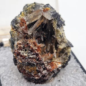 Puede incluir: Un grupo de cristales de cuarzo natural con un exterior oscuro y rugoso. El interior revela numerosos cristales de cuarzo claros y puntiagudos, con toques de depósitos minerales naranjas y marrones. La muestra se presenta sobre un fondo gris claro.