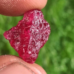 May include: A raw, unpolished ruby gemstone with a deep, vibrant red color and a naturally textured, crystalline surface. The stone features irregular, jagged edges and a translucent quality that allows light to pass through its faceted, uneven structure.