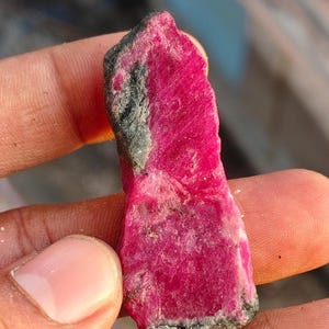 May include: A raw, elongated ruby crystal with a vibrant, deep pinkish-red hue. The stone features a rough, uneven texture with visible crystalline facets and patches of dark, greyish-black mineral matrix along the edges and base.
