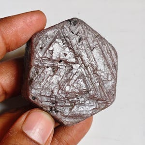 Könnte beinhalten: Ein sechseckiger, roher, bräunlich-grauer Korundkristall mit ausgeprägten dreieckigen Wachstumsmustern und geätzten Oberflächenstrukturen. Das Mineralexemplar zeigt eine geometrische, geschichtete Struktur mit sichtbaren dunklen Einschlüssen auf seiner matten, kristallinen Oberfläche.
