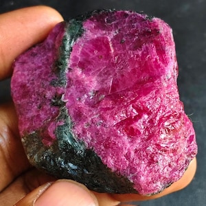 May include: A raw, unpolished ruby gemstone featuring a vibrant, deep magenta-pink crystalline structure with prominent dark, opaque mineral inclusions along the side, showcasing a natural, jagged texture and a translucent, reflective surface.