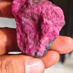 May include: A raw, unpolished ruby gemstone with a vibrant magenta-pink hue and a rough, crystalline texture. The stone features irregular edges and dark grey mineral inclusions embedded within its jagged, natural surface structure.