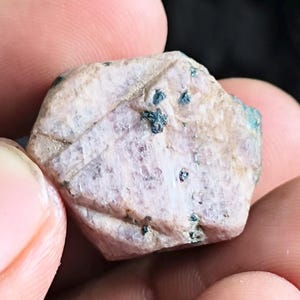 Puede incluir: Una gema en bruto, sin cortar, sostenida en una mano. La piedra es de color rosa pálido con inclusiones azul-verdosas más oscuras. La superficie tiene un aspecto ligeramente texturizado. La palabra "Photographer" está escrita en cursiva en la parte inferior.