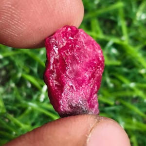 May include: A raw, unpolished ruby gemstone with a deep, vibrant magenta-red colour. The stone features a natural, irregular, and textured surface with visible crystalline facets and a slightly darker, opaque base section at the bottom edge.