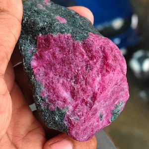Könnte beinhalten: Ein rohes, unpoliertes Mineralexemplar mit einem großen, leuchtend magentafarbenen kristallinen Bereich, der in eine dunkle, texturierte, anthrazitfarbene Gesteinsmatrix eingebettet ist. Der Stein zeigt eine raue, natürliche Oberfläche.