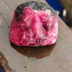 Peut inclure: Un spécimen minéral brut et non poli présentant une structure cristalline rose vif et profonde, contrastée par une matrice texturée gris foncé à noire sur le dessus. La pierre a une forme organique irrégulière avec des facettes visibles.
