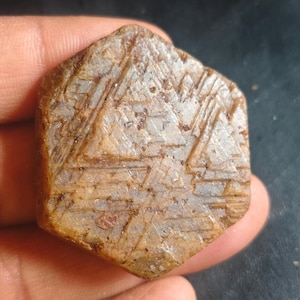 Könnte beinhalten: Ein sechseckiger, roh geschnittener Edelstein in Braun. Die Oberfläche weist geätzte geometrische Muster auf, die ein strukturiertes Aussehen erzeugen. Der Stein wird in einer Hand gehalten, was seine natürliche Form und sein einzigartiges Design hervorhebt.