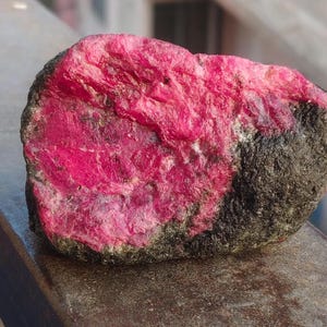May include: A raw, unpolished mineral specimen featuring a vibrant, deep magenta-pink crystalline section embedded within a rough, dark gray, and textured rocky matrix. The stone displays a stark contrast between the bright, faceted-looking pink mineral and the dull, earthy exterior of the surrounding rock formation.