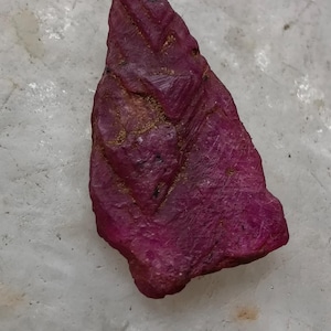 May include: A rough-cut, deep red ruby gemstone with a textured surface. The ruby has a triangular shape and displays natural patterns and variations in color. The gemstone is set against a light-colored background.