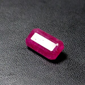 May include: A rectangular, faceted ruby gemstone with a deep pinkish-red hue. The stone features a prominent, flat, reflective white rectangular facet on its top surface, showcasing clear cut edges and a polished, translucent appearance.