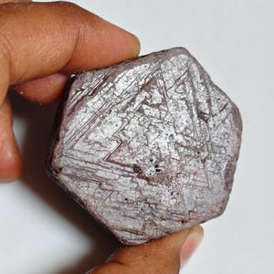 May include: A hexagonal, raw, purplish-gray gemstone featuring a distinct triangular geometric pattern etched across its flat surface. The stone has a matte, textured appearance with visible crystalline striations and small dark inclusions.