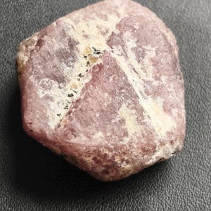 Puede incluir: Una gema rosa rugosa con inclusiones blancas y negras. La piedra tiene una forma irregular y una superficie ligeramente texturizada. La gema se encuentra sobre un fondo gris oscuro.