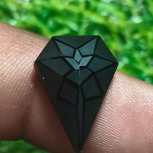 Könnte beinhalten: Ein mattschwarzer, schildförmiger Edelstein mit einer facettierten Oberfläche und einem eingravierten geometrischen, sternartigen Muster auf der Oberseite. Der Stein hat eine spitz zulaufende Unterseite und eine flache, breite Oberkante.