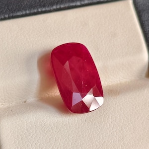 May include: A vibrant, deep red gemstone with a cushion-cut shape, featuring multiple facets that reflect light. The stone displays a rich, saturated crimson hue and is positioned on a textured, off-white surface with a dark border.