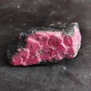 Peut inclure: Un spécimen minéral brut et non poli présentant une section cristalline magenta-rose vive et saturée, contrastée par une croûte extérieure rugueuse d'un noir charbon foncé. La pierre a une forme irrégulière et allongée.