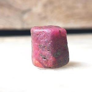 Peut inclure: Une pierre précieuse rubis brute de forme cylindrique avec une teinte magenta et rose profonde. La surface de la pierre est texturée et opaque, présentant des inclusions naturelles et des dépôts minéraux sombres sur tout son extérieur.