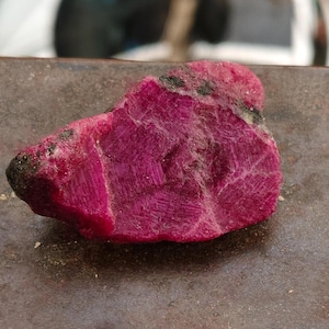 May include: A raw, unpolished ruby gemstone with a deep magenta-red color. The stone features a rough, crystalline texture with visible geometric striations and small dark mineral inclusions scattered across its uneven, jagged surface edges.