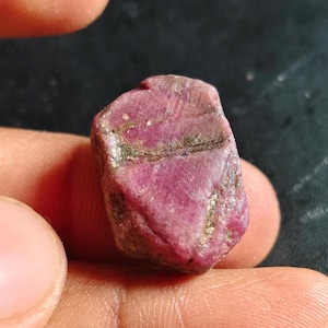 Può includere: Una gemma di rubino grezza, non tagliata, con una tonalità rosso-rosata intensa. La superficie presenta texture naturali e alcune inclusioni più scure, marrone-verdastre. La gemma è tenuta in mano, mostrando la sua bellezza naturale. Una gemma naturale.