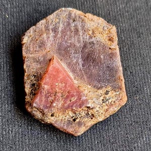 Puede incluir: Una gema de rubí natural, tallada en bruto con forma hexagonal. El rubí presenta una inclusión triangular de color rojo intenso dentro de un cuerpo translúcido de color rojo púrpura. Los bordes son marrones y dorados, con una superficie texturizada.