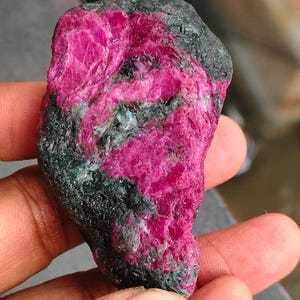 以下が含まれることがあります： 鮮やかな深いマゼンタピンクの結晶中心部を持つ、未研磨の原石標本。縁や結晶構造の内部には、暗い質感のチャコールグレーから黒色の母岩が混ざり合い、鮮やかなピンク色とのコントラストを生み出しています。