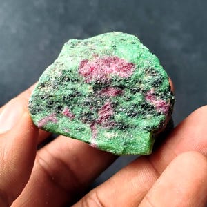 Puede incluir: Una gema en bruto con una vibrante combinación de colores. La piedra muestra una llamativa mezcla de tonos rojos y rosas intensos, intercalados con áreas de verde intenso. La textura general parece áspera y natural, con formaciones minerales visibles.