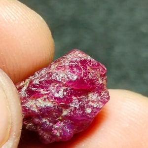 May include: A raw, unpolished ruby gemstone with a deep, saturated magenta-red colour. The stone features a natural, irregular crystalline structure with visible facets, textured surfaces, and translucent areas that catch the light.