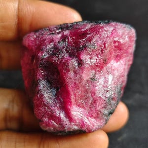 May include: A raw, unpolished ruby gemstone with a deep magenta and crimson colour palette. The stone features a rough, textured surface with visible crystalline structures and dark, opaque mineral inclusions throughout its irregular shape.