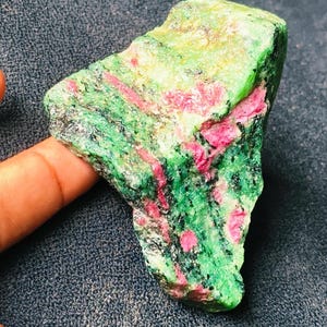 May include: A rough-cut gemstone with a vibrant mix of colors. The stone features a base of green with streaks and patches of bright pink and yellow. The texture appears rough and natural, with visible mineral formations.