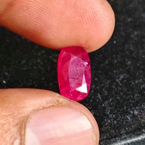 May include: A vibrant, oval-cut gemstone with a deep red color and multiple facets that reflect light. The stone displays a translucent quality with visible internal inclusions and a polished surface, showcasing a brilliant, saturated crimson hue.