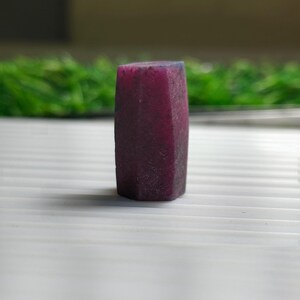 May include: A raw, hexagonal-shaped gemstone with a deep magenta and dark red colour. The stone features a matte, textured surface with visible natural facets and a vertical orientation, standing upright on a flat, light-coloured surface.