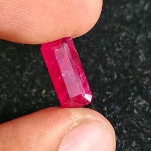 May include: A vibrant, rectangular-cut ruby gemstone with a deep red hue and multiple facets that reflect light. The stone is held between two fingers, showcasing its transparent quality and precise geometric shape against a dark background.