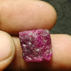 May include: A raw, unpolished ruby gemstone with a deep magenta-red colour and a rough, textured surface. The stone has an irregular, somewhat rectangular shape with visible natural inclusions and crystalline facets across its surface.