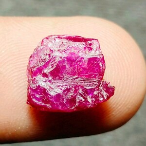 May include: A raw, unpolished ruby gemstone with a vibrant, deep pinkish-red colour. The stone features a natural, irregular, and textured surface with visible crystalline facets and edges, showcasing its authentic, untreated, and rough mineral form.