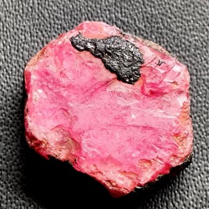 Peut inclure: Une pierre précieuse rose brute avec une inclusion sombre et texturée. La pierre a une couleur vive et saturée et une forme légèrement irrégulière. La surface présente des variations et des textures naturelles, suggérant qu'elle est non polie. Il s'agit probablement d'un spécimen minéral brut.