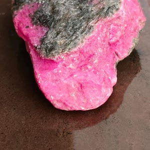 Puede incluir: Un espécimen mineral en bruto y sin pulir que presenta una sección de color rosa intenso saturado que contrasta con una formación cristalina de color gris carbón oscuro y texturizado. La piedra tiene una forma irregular y dentada.
