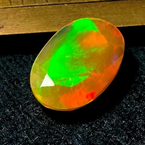 May include: An oval-cut opal gemstone displaying vibrant flashes of neon green and orange play-of-color. The gemstone is positioned next to a metal ruler, showing a portion of the scale with markings visible at the top edge of the frame.