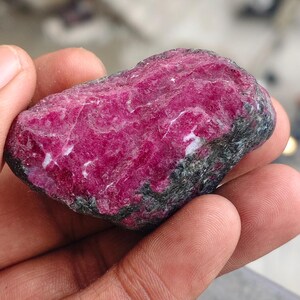 May include: A raw, unpolished gemstone featuring a vibrant, deep magenta-pink crystalline surface with visible white mineral veins and dark, rough, charcoal-colored rock matrix along the edges and base of the stone.