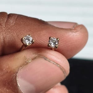 May include: A pair of gold stud earrings, each featuring a clear, round-cut gemstone. The earrings are held between fingers, showcasing the delicate design and sparkle of the jewelry.