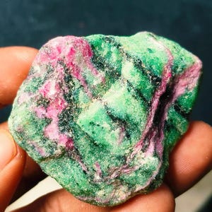 Può includere: Una gemma grezza con colori vivaci. La pietra presenta una sorprendente combinazione di verde intenso e rosa acceso, con una superficie strutturata. Il minerale naturale è tenuto in una mano, mostrando la sua bellezza grezza e non lucidata.