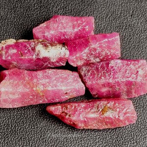 May include: A collection of rough-cut, raw ruby gemstones. The rubies are a vibrant pinkish-red color, with a slightly textured surface. They are arranged on a dark gray, textured background.