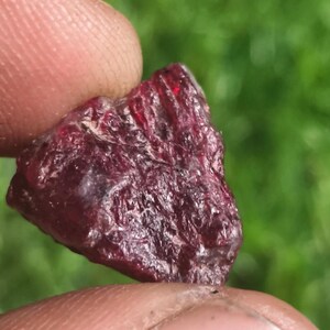 May include: A raw, deep red gemstone with a textured, crystalline surface. The stone displays irregular facets and natural inclusions, showing a dark crimson hue that catches light in specific areas, revealing translucent properties within the structure.