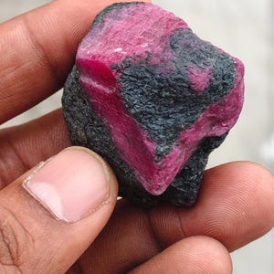 May include: A raw, unpolished gemstone featuring a vibrant, deep pinkish-red crystalline structure embedded within a dark, textured, charcoal-colored matrix. The stone displays a jagged, irregular shape with contrasting mineral layers.