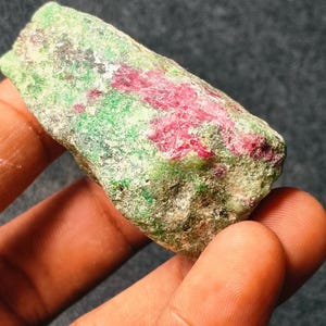 May include: A raw gemstone displaying a blend of green and red colours. The stone has a textured surface, suggesting a natural mineral. The red is streaked across a green base, creating a striking visual contrast.
