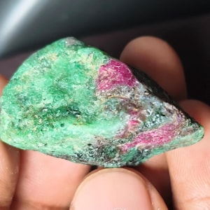 Può includere: Una gemma grezza con una miscela di tonalità verdi e rosse. La pietra ha una superficie strutturata, con aree di verde scuro e macchie di rosso vibrante. La forma generale è irregolare, mostrando la sua forma naturale.