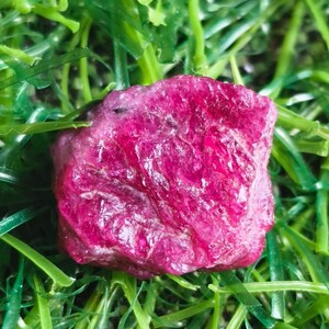 May include: A raw, unpolished ruby gemstone with a deep magenta-pink hue and a textured, crystalline surface. The stone features irregular facets and natural inclusions, displaying a vibrant, saturated colour throughout its jagged, organic shape.