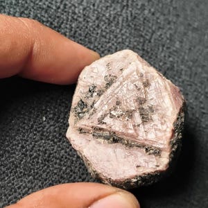 Puede incluir: Una gema rugosa, de color blanco rosado, con inclusiones oscuras y una estructura cristalina. La piedra se sostiene entre los dedos, mostrando su aspecto natural y sin pulir. La roca tiene una superficie rugosa y texturizada.