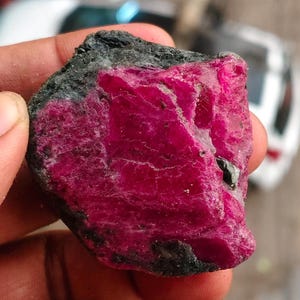 Könnte beinhalten: Ein roher, ungeschliffener Edelstein mit einem leuchtenden, tief magentafarbenen kristallinen Kern, der in eine raue, dunkelgraue bis schwarze Mineralmatrix eingebettet ist. Der Stein hat eine unregelmäßige, gezackte Form mit sichtbaren Texturen.